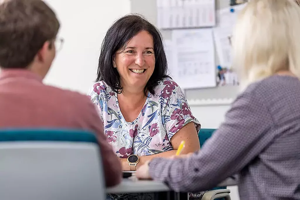 Gespräch Eine Mitarbeiterin unterhält sich mit zwei Patienten am Schreibtisch.