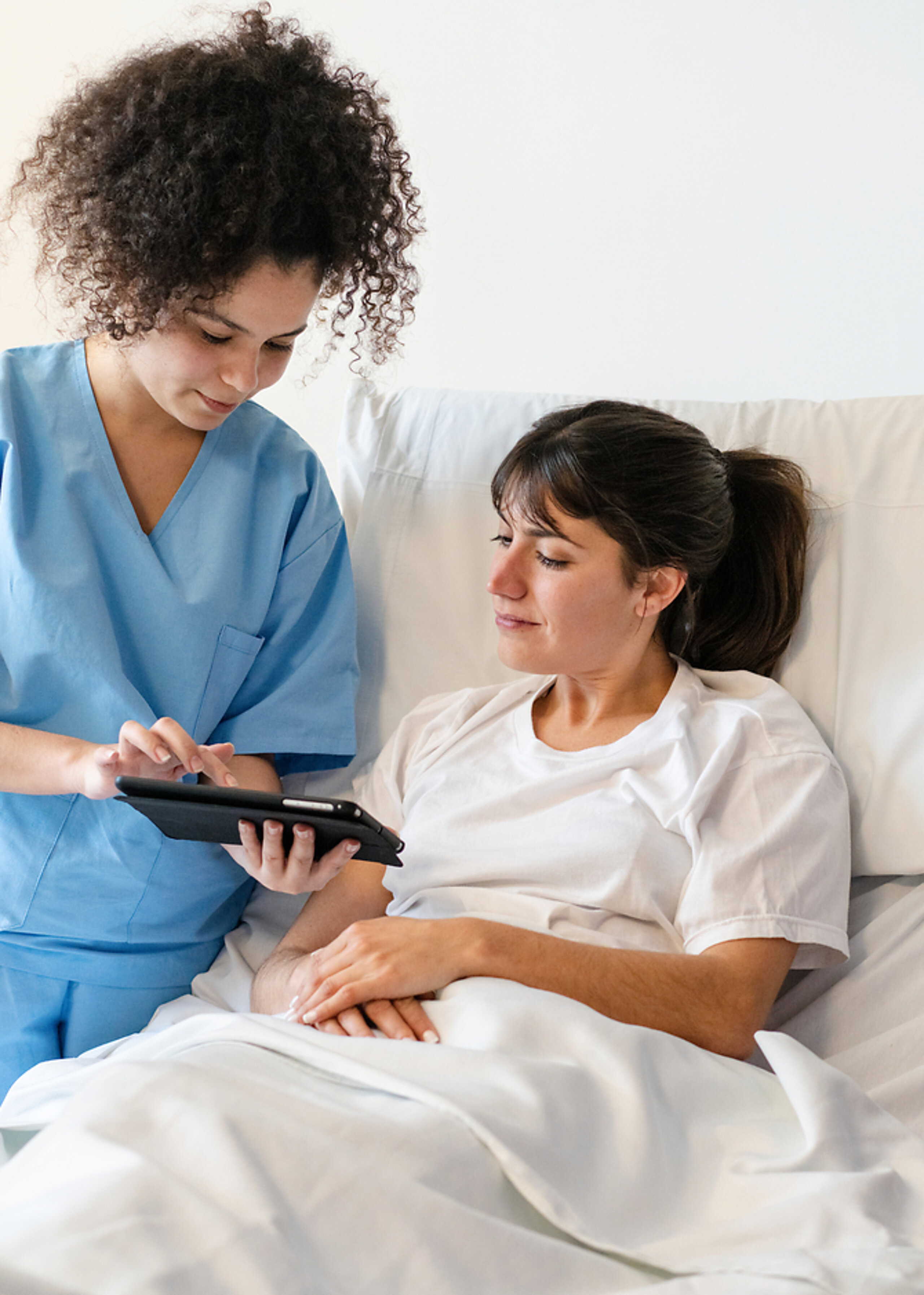 Pflege digital Eine Pflegerin zeigt einer Patientin im Krankenhausbett etwas auf dem Tablet.