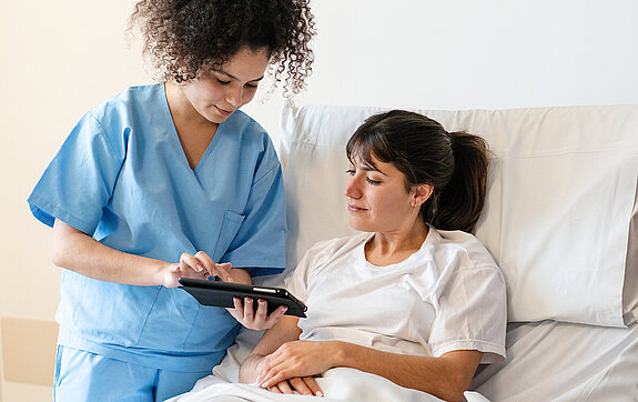 Pflege digital Eine Pflegerin zeigt einer Patientin im Krankenhausbett etwas auf dem Tablet.