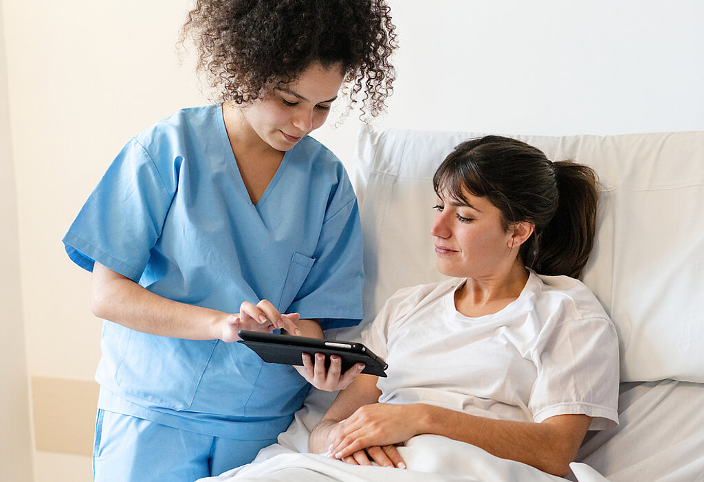 Pflege digital Eine Pflegerin zeigt einer Patientin im Krankenhausbett etwas auf dem Tablet.