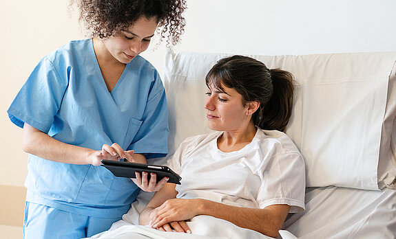 Pflege digital Eine Pflegerin zeigt einer Patientin im Krankenhausbett etwas auf dem Tablet.