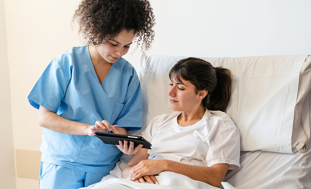 Pflege digital Eine Pflegerin zeigt einer Patientin im Krankenhausbett etwas auf dem Tablet.
