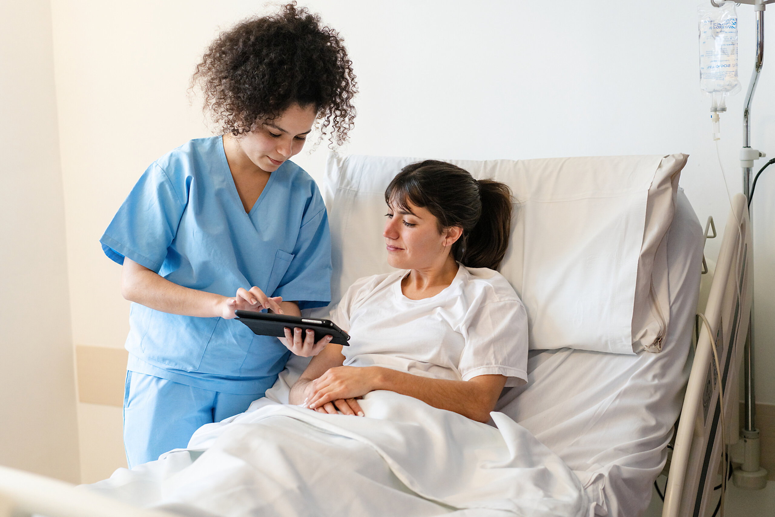 Pflege digital Eine Pflegerin zeigt einer Patientin im Krankenhausbett etwas auf dem Tablet.