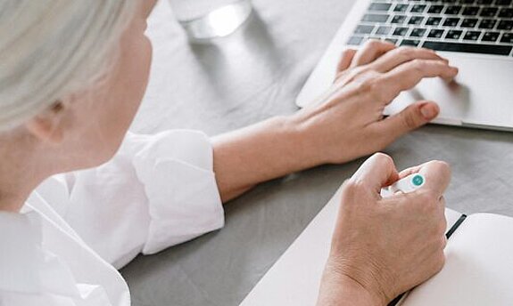 Frau am Laptop Eine ältere Frau sucht etwas am Laptop und macht sich dabei Notizen auf einen Block.