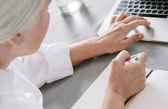 Frau am Laptop Eine ältere Frau sucht etwas am Laptop und macht sich dabei Notizen auf einen Block.